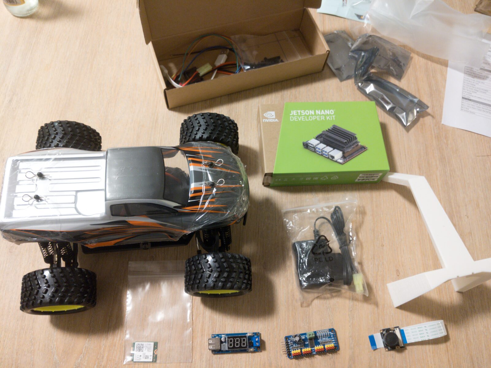 DonkeyCar parts laid out on a wooden floor — an RC monster-truck chassis with bodyshell, an NVIDIA Jetson Nano Developer Kit box, a PCA9685 PWM driver board, a buck converter, a CSI camera module and a white 3D-printed mounting cage