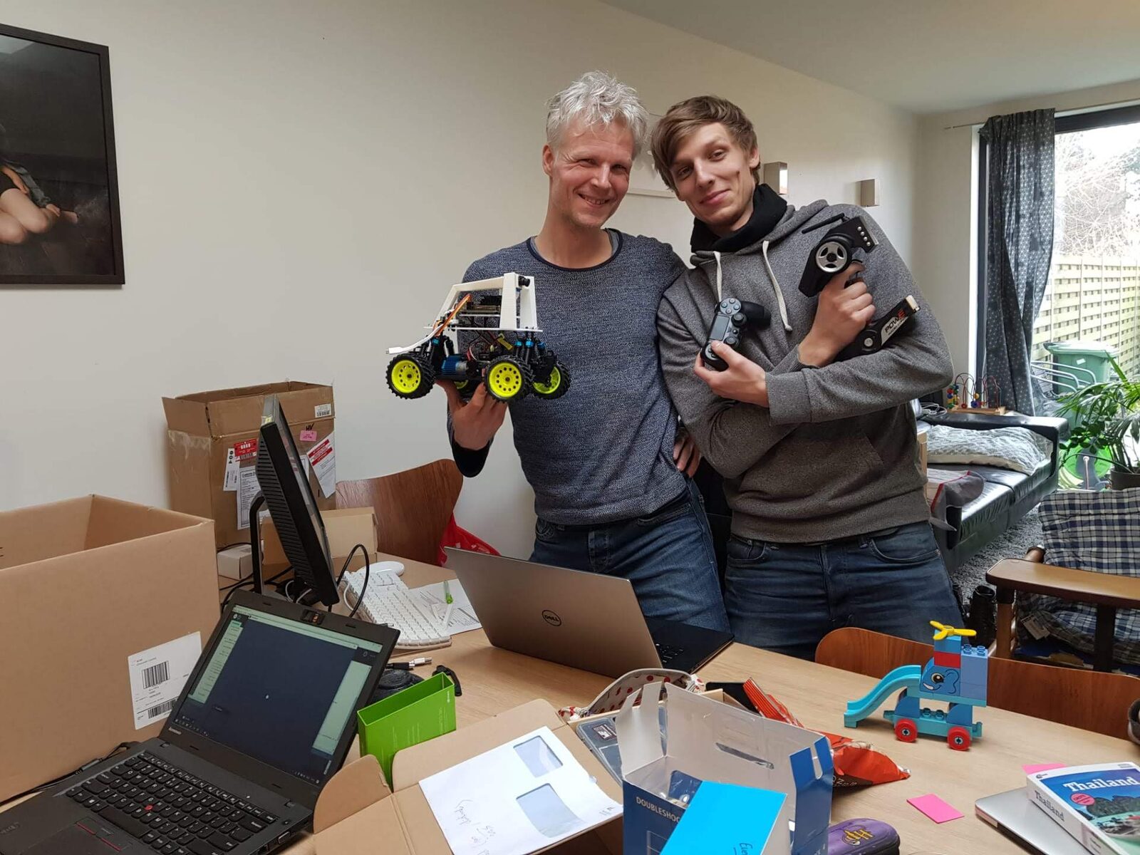 Two people smiling and holding up the assembled DonkeyCar and a black DualShock-style controller; a blue "DOUBLESHOCK" retail box sits on the cluttered desk in front of them
