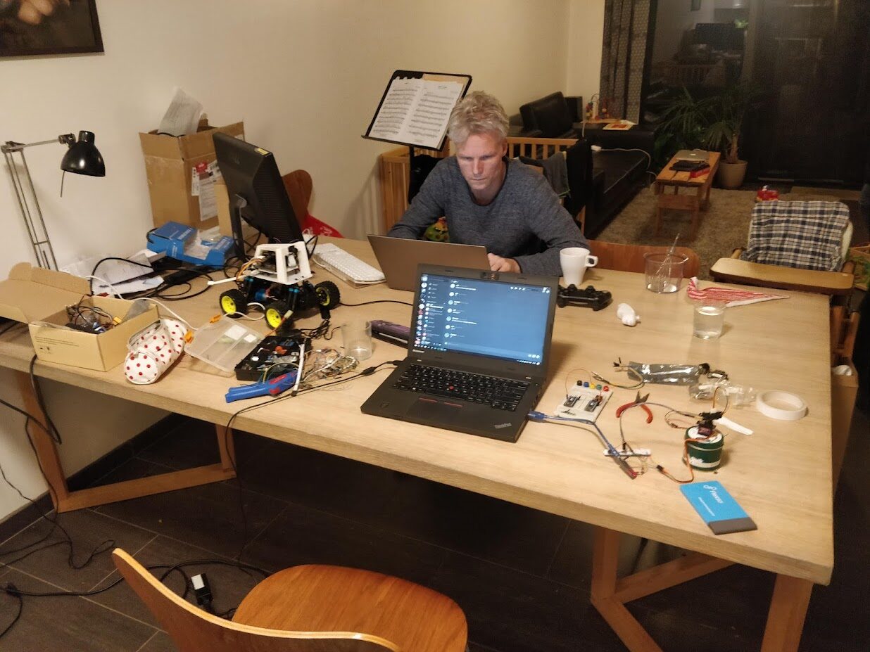A person working at a laptop late at night at a wooden dining table cluttered with the DonkeyCar, a breadboard, components and a DualShock controller